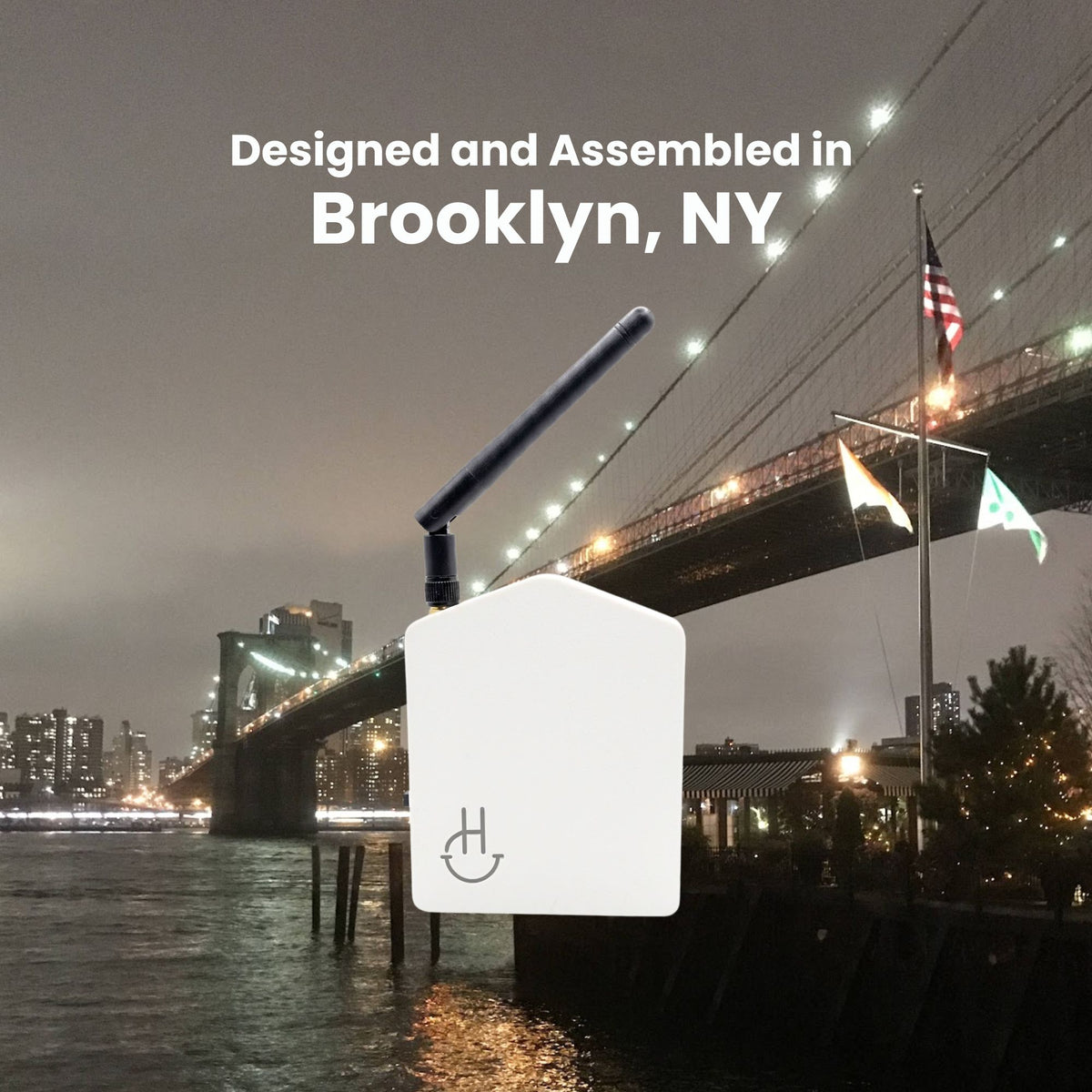 A white rectangular electronic device with a black antenna and a power symbol on the front, shown against a nighttime city waterfront background with the Brooklyn Bridge and text reading 'Designed, Assembled in Brooklyn, NY'.