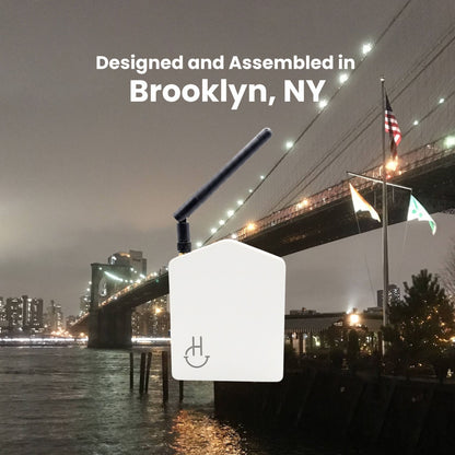A white rectangular electronic device with a black antenna and a power symbol on the front, shown against a nighttime city waterfront background with the Brooklyn Bridge and text reading 'Designed, Assembled in Brooklyn, NY'.